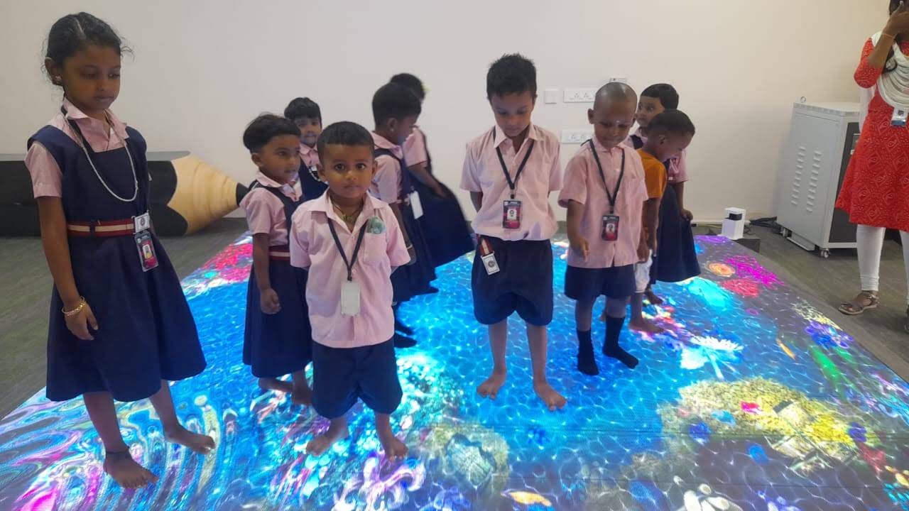 Barefoot children in uniform standing on a glowing, colourful interactive floor projection
