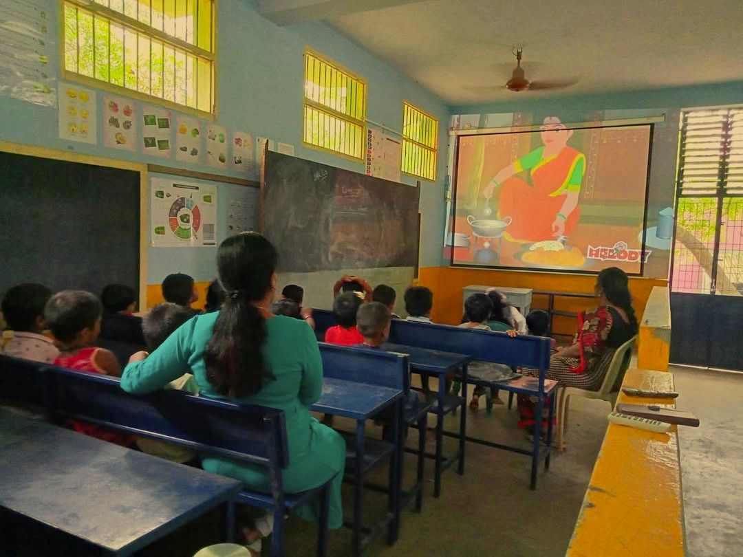 Children and teachers on classroom benches watching an animated video on a large projector screen